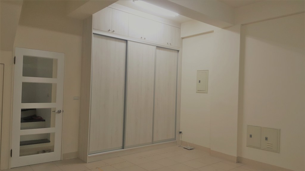 一間簡約的室內空間，牆上有帶滑門的衣櫃，旁邊是一扇白色的玻璃門，地板是淺色瓷磚。